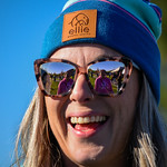 A woman wearing a blue beanie with a leather patch and sunglasses smiles brightly.