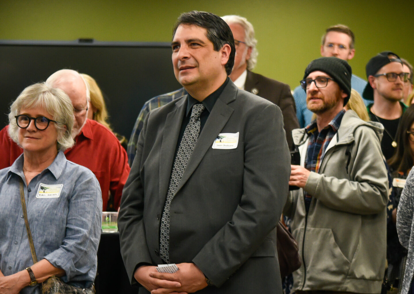 A group of people attends an indoor professional event, listening attentively. The central figure is a man in a dark suit with a name tag, surrounded by attendees in both formal and casual attire.