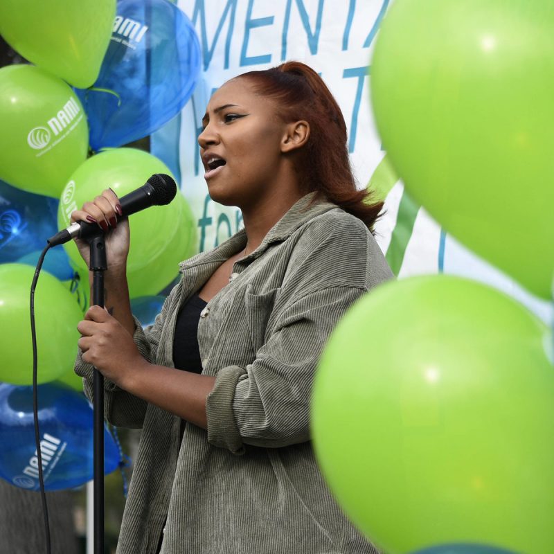 A young black woman speaking at a NAMI event with balloons in the background